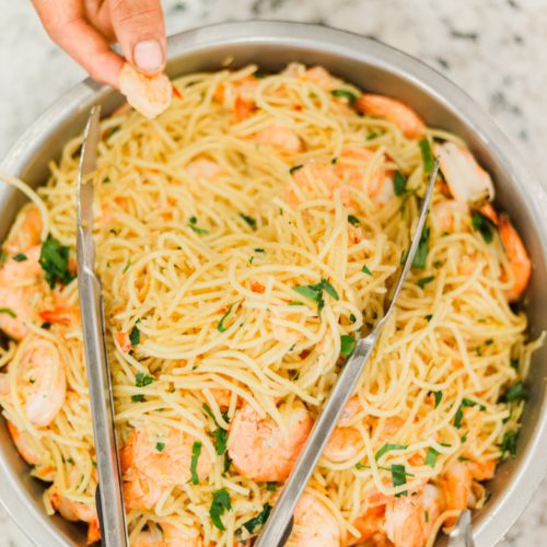angel hair pasta mixed with shrimp and green onions in a silver metal bowl with tongs